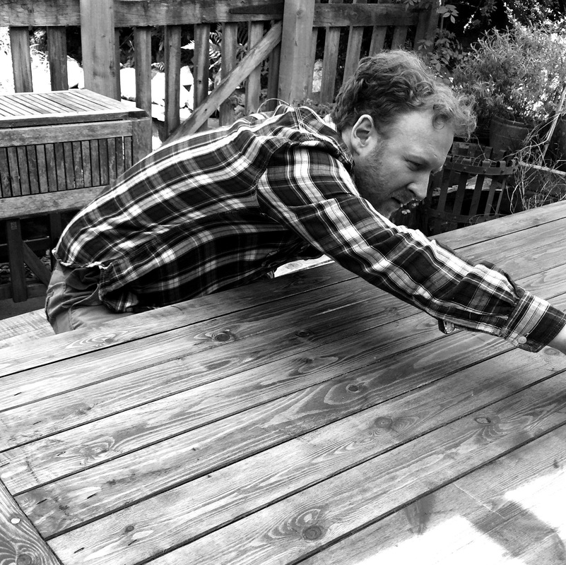 A black and white photo of a man wearing a checked shirt, sanding a wooden table.